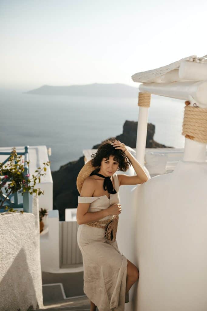 Brunette lady in beige midi dress touches hair. Curly charming woman in stylish outfit with straw b